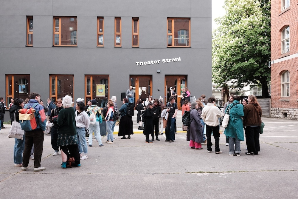Theater von außen. Menschen sammeln sich und warten vor dem Gebäude auf die Vorstellung.