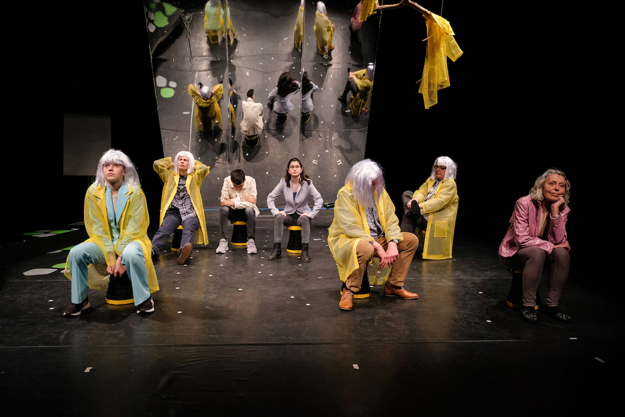 7 sitzende Personen in Kostümen auf der Bühne.